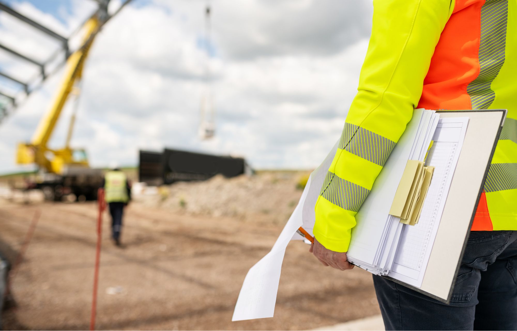Widok od tyłu na osobę w żółtej kamizelce, która trzyma plik dokumentów. W tle plac budowy.